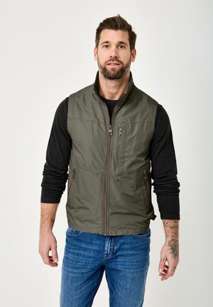 Mann mit Bart, der eine olivgrüne Zip-Up-Weste über einem schwarzen Langarmshirt und blauen Jeans trägt, steht mit entspannten Armen.