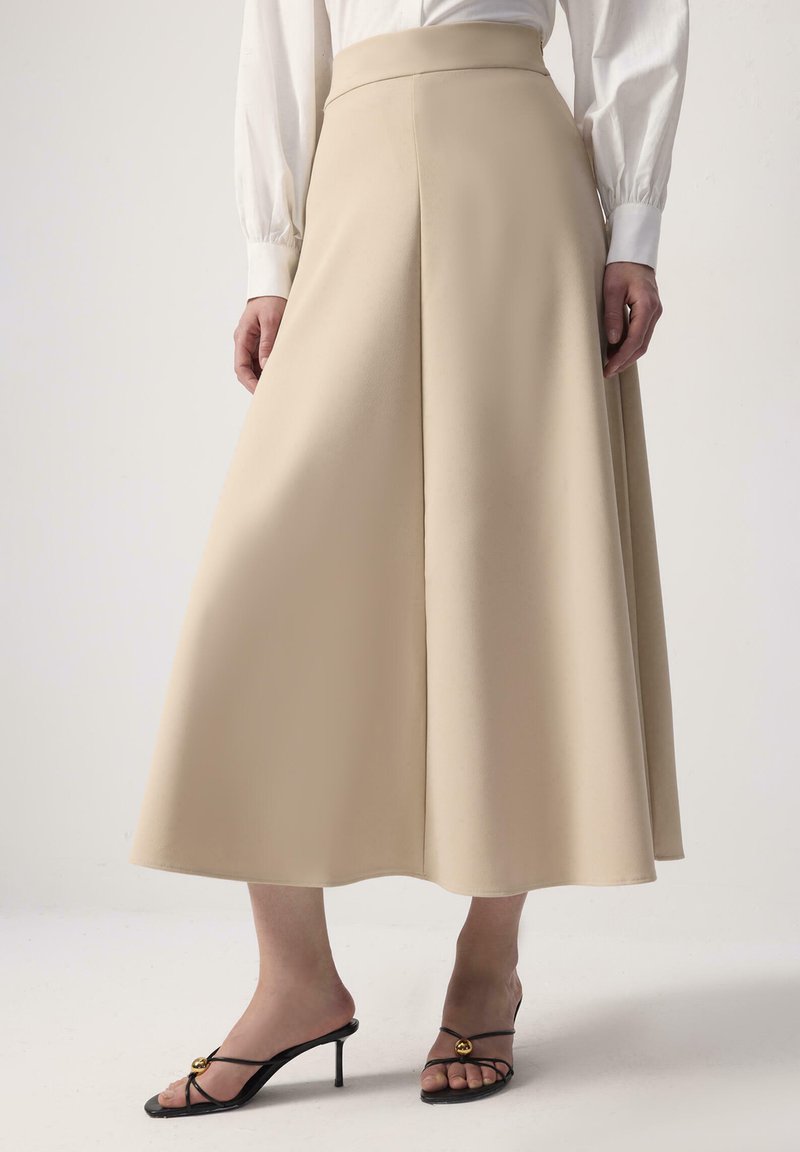 Falda beige hasta la rodilla con silueta en A, líneas limpias y una cinturilla estructurada, combinada con tacones negros con tiras y acentos dorados.