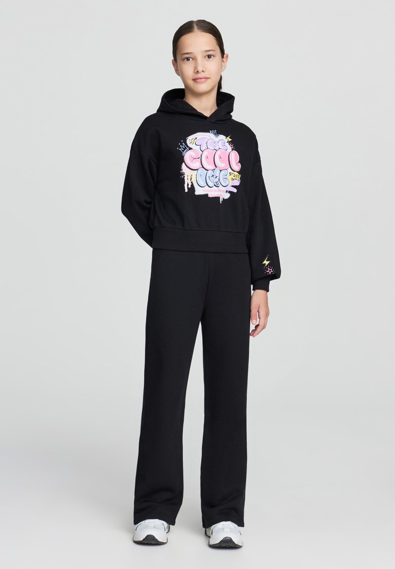 Felpa nera corta con una stampa grafica colorata, abbinata a pantaloni neri a gamba larga e sneakers bianche. Tessuto liscio con una vestibilità comoda.