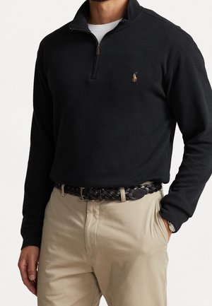 Man wearing black quarter-zip pullover with orange embroidered logo, beige pants, black braided belt, white undershirt, watch, and hand in pocket.