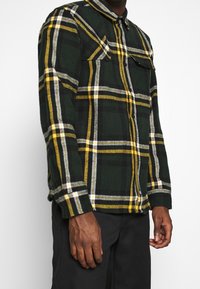 Camicia di flanella verde con motivo a quadri con accenti gialli e bianchi, due tasche sul petto e colletto con bottoni. Tessuto testurizzato.