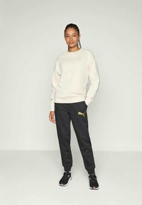 Beige kapucnis pulóver bordás mandzsettákkal és aljjal, fekete melegítőnadrággal párosítva, arany logóval. Fekete-fehér sportcipők teszik teljessé az outfitet.