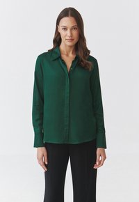 Blusa di raso verde con colletto e chiusura a bottoni, caratterizzata da maniche lunghe e una vestibilità comoda, abbinata a pantaloni neri.