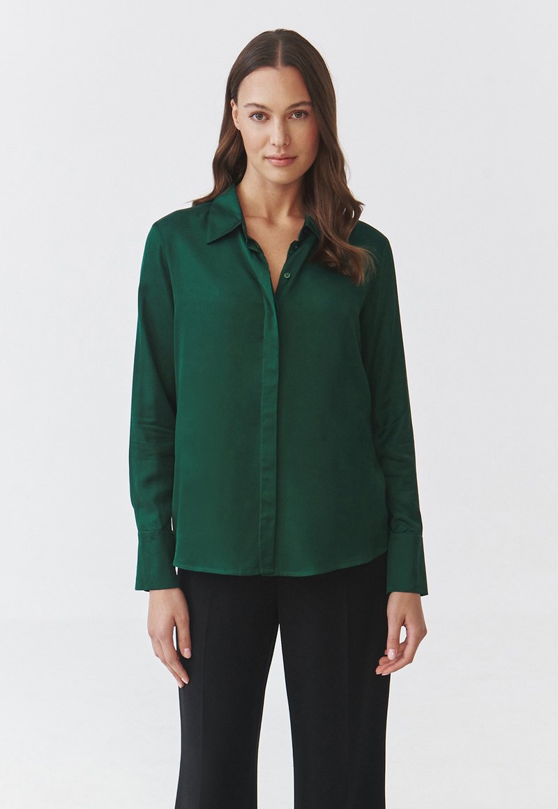 Blusa di raso verde con colletto e chiusura a bottoni, caratterizzata da maniche lunghe e una vestibilità comoda, abbinata a pantaloni neri.