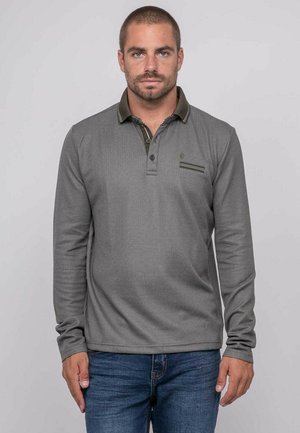 Homme aux cheveux courts et barbe portant un polo gris à manches longues et un jean bleu, debout devant un fond uni.