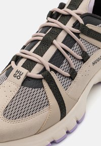 Beige sportskor med grå mesh, svarta läderdetaljer och mönstrade snören. Har en lätt sula med lila spets och logodetalj.