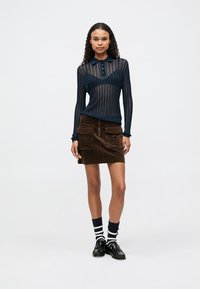 Haut en tricot bleu marine à manches longues avec des rayures discrètes, accompagné d'une jupe en velours côtelé marron avec deux poches avant et des chaussures à lacets foncées.