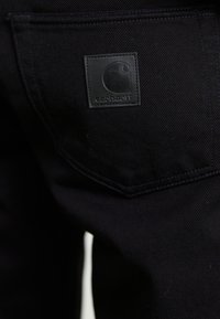 Zwarte denim broek met een gestikte achterzak en een textuurgepatch van zwart leer waarop een logo en merknnaam staan.
