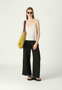 Top alb cu detalii decorative aurii, pantaloni negri cu croială largă, geantă supradimensionată galbenă și sandale negre flip-flop, completate cu ochelari de soare închiși la culoare.