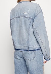 Světle modrá džínsová bunda s volným střihem, s poškozenými detaily, viditelným šitím a třepeným lemem na pásku.