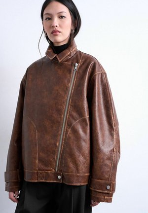 Joven mujer con chaqueta de cuero marrón extragrande con cremallera diagonal, de pie frente a un fondo claro liso.