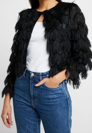 Femme portant une veste noire texturée à franges sur un haut blanc et un jean taille haute bleu, une main dans la poche, cadrée au niveau du cou.