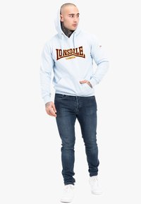 Felpa hoodie azzurro chiaro con coulisse e un grande logo bordeaux "Lonsdale London", abbinata a jeans di denim scuri e sneaker bianche.