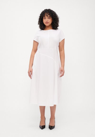 Robe midi blanche à manches courtes, col rond et taille structurée. Elle présente un détail de couture sur le côté et est associée à des talons aiguilles noirs.