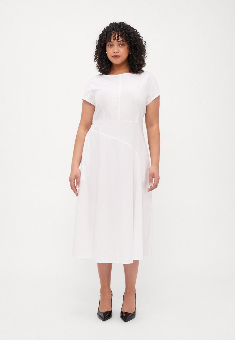 Robe midi blanche à manches courtes, col rond et taille structurée. Elle présente un détail de couture sur le côté et est associée à des talons aiguilles noirs.