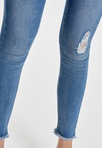Jeans ajustados de mezclilla azul con detalles desgastados, incluyendo un área rasgada en el muslo y dobladillos deshilachados en el tobillo.