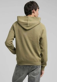 Felpa con cappuccio verde oliva dal tessuto liscio, cappuccio con coulisse e polsini a coste. Indossata con jeans grigio chiaro; visualizzata da dietro.