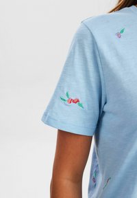Camiseta de manga corta azul claro con bordado floral en las mangas, que presenta flores rosas y hojas verdes en una tela suave.