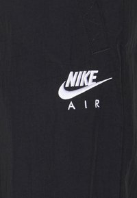 Černý textilní oděv s bílou vyšívanou logem "NIKE" nad slovem "AIR". Textura vypadá mírně pomačkaně s viditelnými detaily šití.