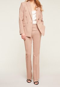 Světle růžový třpytivý oblek skládající se z dvouřadého saka s klopami a přizpůsobených rozšířených kalhot. Nosit s bílým topem a černými sandály.