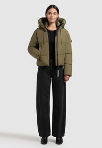 Olive green puffer jacket with a hood, featuring a zip front, two side pockets, and a rectangular quilted design. Worn with black wide-leg pants.