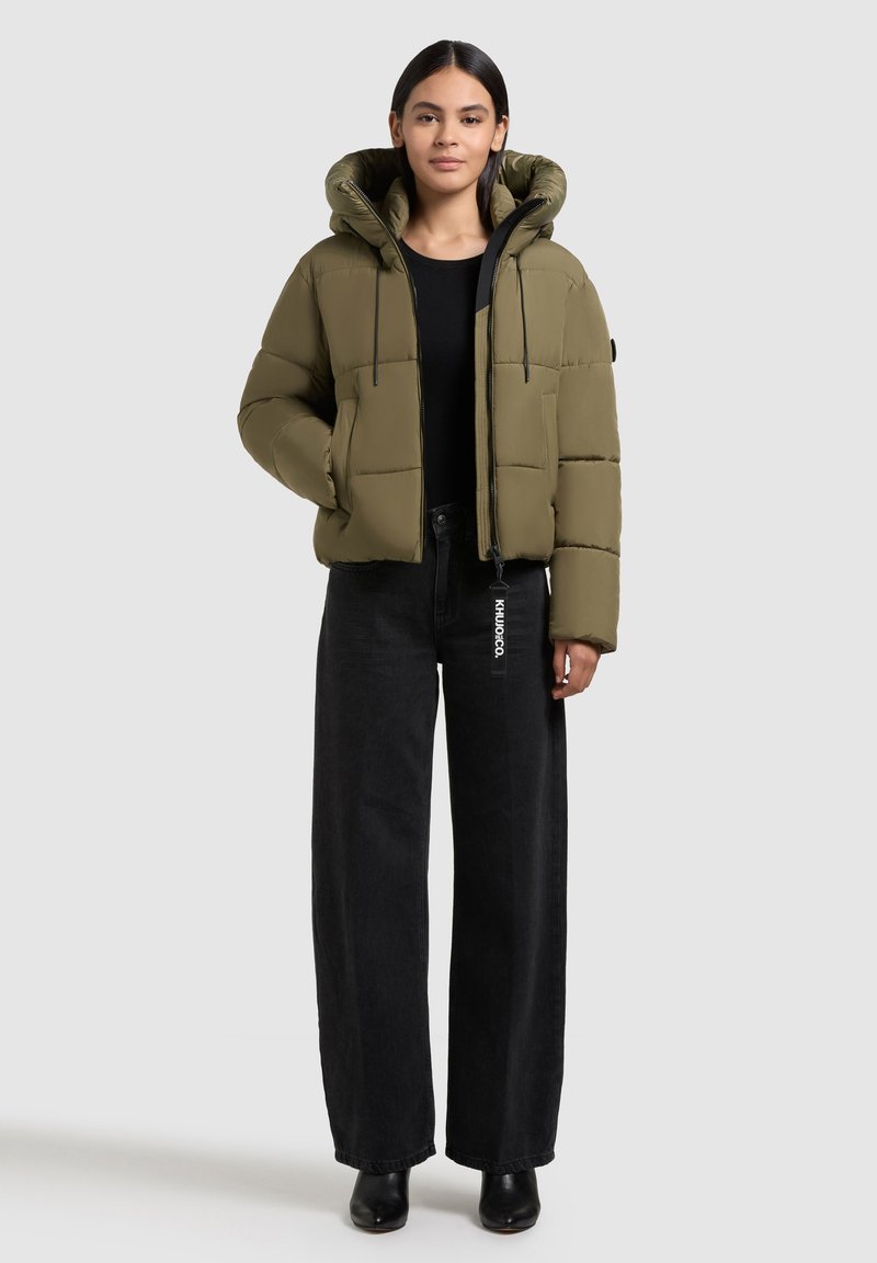 Chaqueta de plumas color verde oliva con capucha, con cierre de cremallera en el frente, dos bolsillos laterales y un diseño acolchado rectangular. Combinada con pantalones anchos negros.