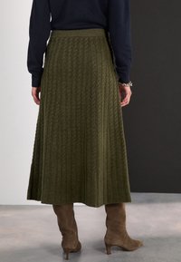 Personne portant une jupe midi en maille torsadée vert olive, un haut à manches longues bleu marine, un bracelet argenté et des bottines en daim marron à talons, debout sur un sol poli.