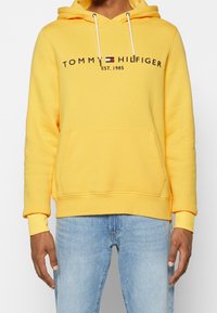 Sudadera amarilla con capucha, cordones y bolsillo frontal, con el texto "TOMMY HILFIGER EST. 1985" en negro. Material de algodón, ajuste holgado.
