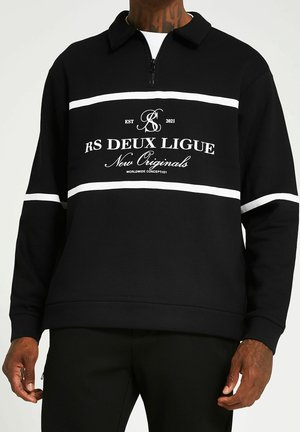 Schwarzer Langarmpullover mit weißen horizontalen Streifen und dem Text „RS DEUX LIGUE New Originals“ auf der Brust, getragen von einer Person mit tätowierten Händen.