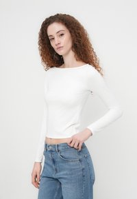 Γυναίκα με σγουρά κόκκινα μαλλιά, φορώντας λευκή εφαρμοστή μπλούζα με μακριά μανίκια και μπλε τζιν, πόζαρε μπροστά σε ένα απλό ανοιχτόφωτο φόντο.