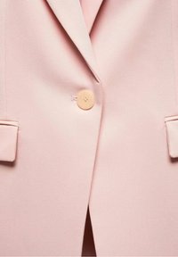 Veste rose à col cranté, un bouton en bois clair, et poches à rabat. Tissu lisse, coupe ajustée avec des lignes épurées.
