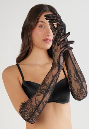 Gants en dentelle noire avec des motifs floraux, manches longues, texture transparente et bords festonnés, portés par-dessus un soutien-gorge en satin noir.