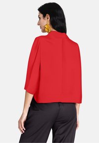 Blusa rossa corta con colletto alto, maniche ampie e una consistenza morbida. Abbinata a pantaloni neri su misura, con orlo dritto.