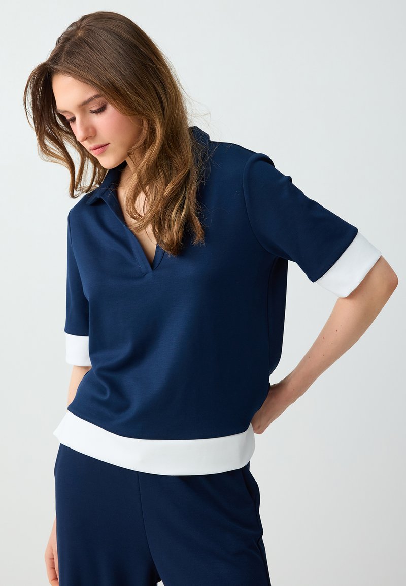 Jeune femme portant une chemise bleu marine à manches courtes avec poignets et ourlet blancs, debout, une main sur la hanche, regardant vers le bas.