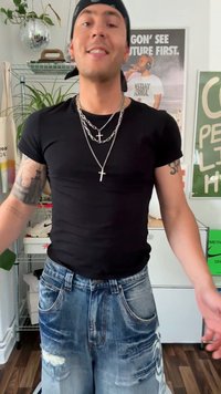 Melnas pieguļošas T-krekls, slāņi sudraba ķēdēm ar krusta kuloniem, nolietotas zilas džinsu bikses ar lieliem plīsumiem. iekštelpu vide ar zaļumiem.