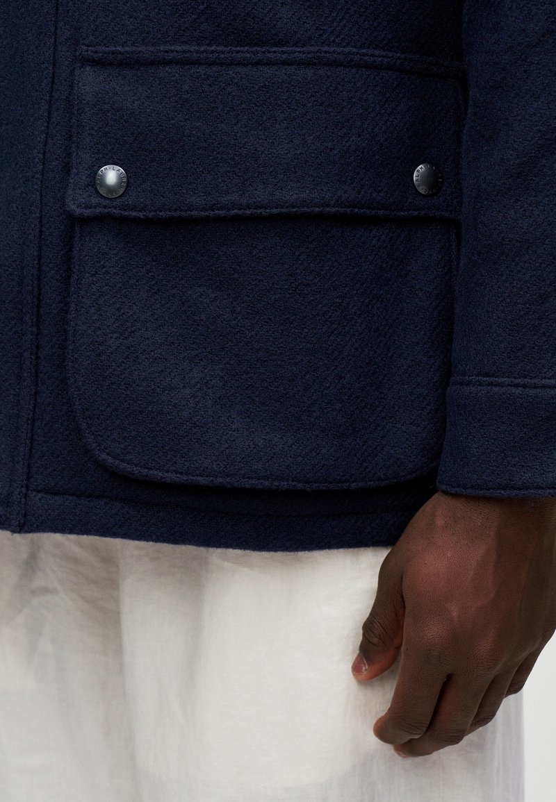 Veste en laine bleu marine foncé avec poche à bouton sur le devant, portée sur un tissu blanc, avec une main à la peau foncée visible reposant sur le côté.