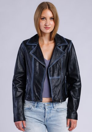 Jeune femme aux cheveux châtain clair et lisses porte une veste en cuir noire, un crop top gris et un jean bleu clair déchiré sur fond gris.