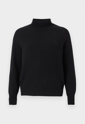 Kobieta ubrana w czarny sweter z wysokim golfem i długimi rękawami, białe szerokie spodnie i czarne buty na obcasie z odkrytymi palcami, stojąca na jednolitym tle.