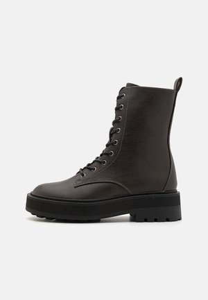 Dunkelbrauner Schnür-Kampfstiefel mit dicker schwarzer Gummisohle und Zuglasche hinten, seitlich auf weißem Hintergrund dargestellt.