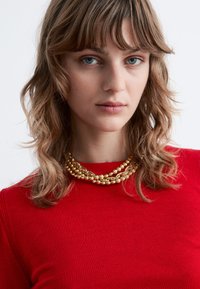 Gold multi-strand necklace with spherical beads worn over a red knit top, featuring a high neckline and textured fabric.