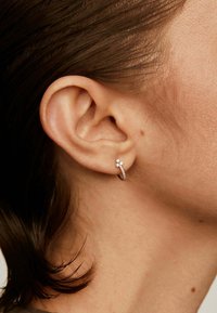 Pendiente de aro de plata con un pequeño diseño de flor y piedras preciosas transparentes, en primer plano de la oreja y el cabello de una persona.
