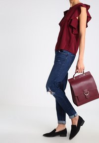 Burgunderfarget top med flæser parret med slidte blå jeans, sorte flade sko og en maroon håndtaske med en guldringe detaljer.