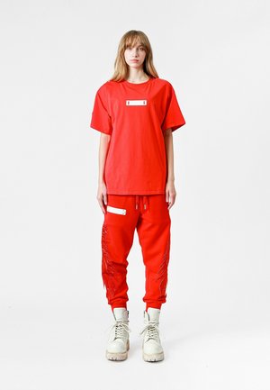 Camiseta de algodón roja con un parche blanco rectangular, combinada con pantalones deportivos rojos que tienen bolsillos laterales y un detalle texturizado. Botas blancas.