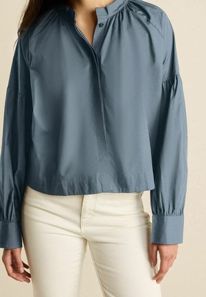 Femme portant un chemisier bleu ardoise à manches longues avec un col montant et un pantalon crème sur un fond uni.