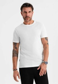 Camiseta blanca de manga corta en suave tejido de algodón, con cuello redondo y forma ajustada, combinada con pantalones negros y un reloj de plata.