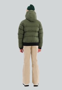 Persoon met een groene beanie en een olijfgroene gewatteerde jas met capuchon, beige broek en zwarte schoenen, staand met de rug naar de camera tegen een grijze achtergrond.