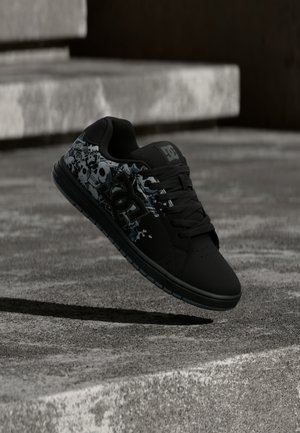 Chaussure de skate noire avec graphisme de crâne et logo DC, suspendue en l'air au-dessus de marches en béton dans un cadre extérieur.