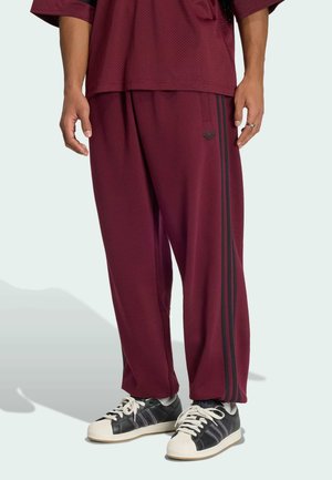 Osoba ubrana w bordowe spodnie dresowe Adidas z czarnymi paskami oraz czarne buty sportowe Adidas z białymi czubkami i sznurówkami.