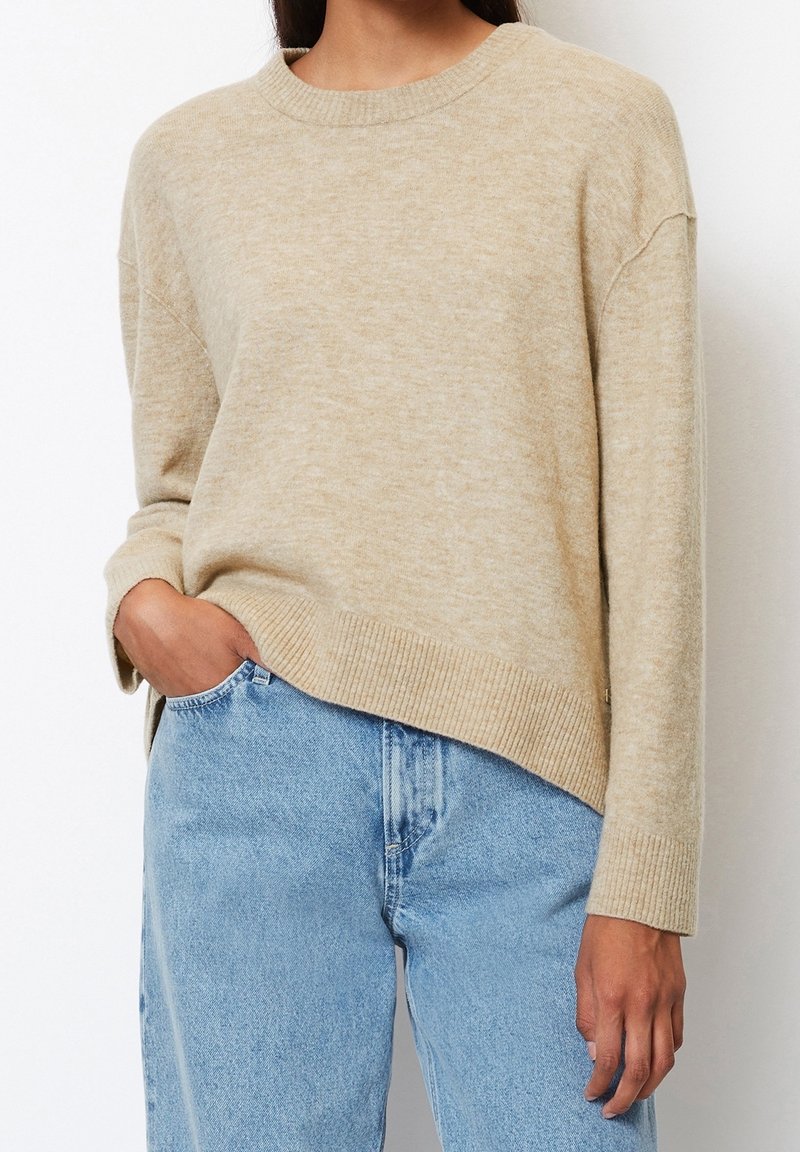 Pull en laine beige avec encolure ronde, poignets et ourlet côtelés. Associé à un jean taille haute bleu clair. Texture douce, coupe décontractée.