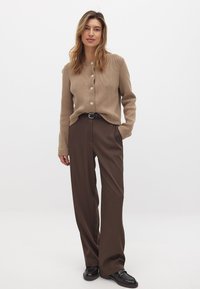 Beige gebreide cardigan met een ronde hals en knopen, gecombineerd met hooggesneden, loszittende donkerbruine broek en zwarte schoenen.